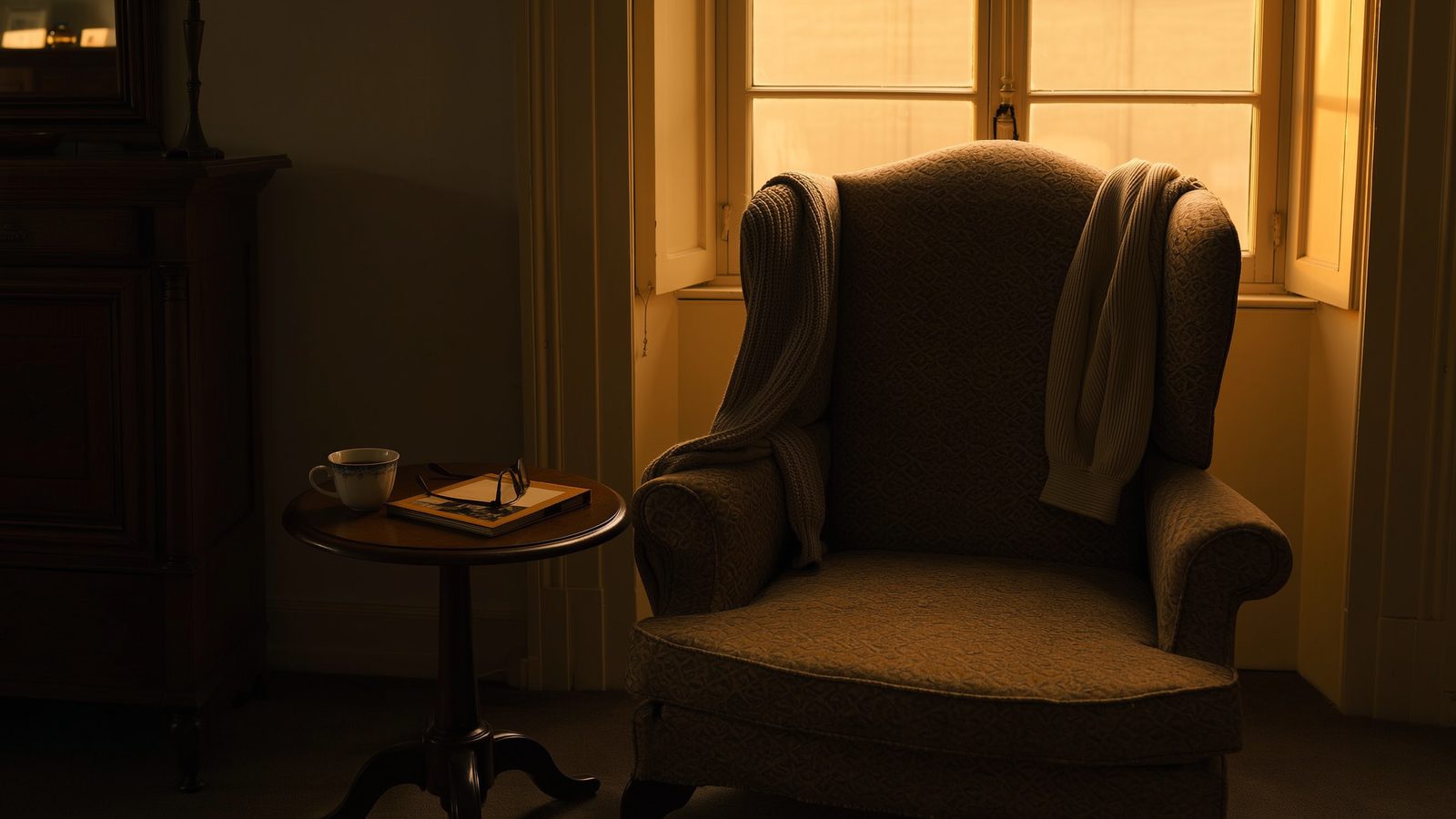 Ein abgewetzter Sessel am Fenster im Abendlicht, eine Strickjacke über der Lehne, daneben ein geschlossenes Fotoalbum mit Lesebrille