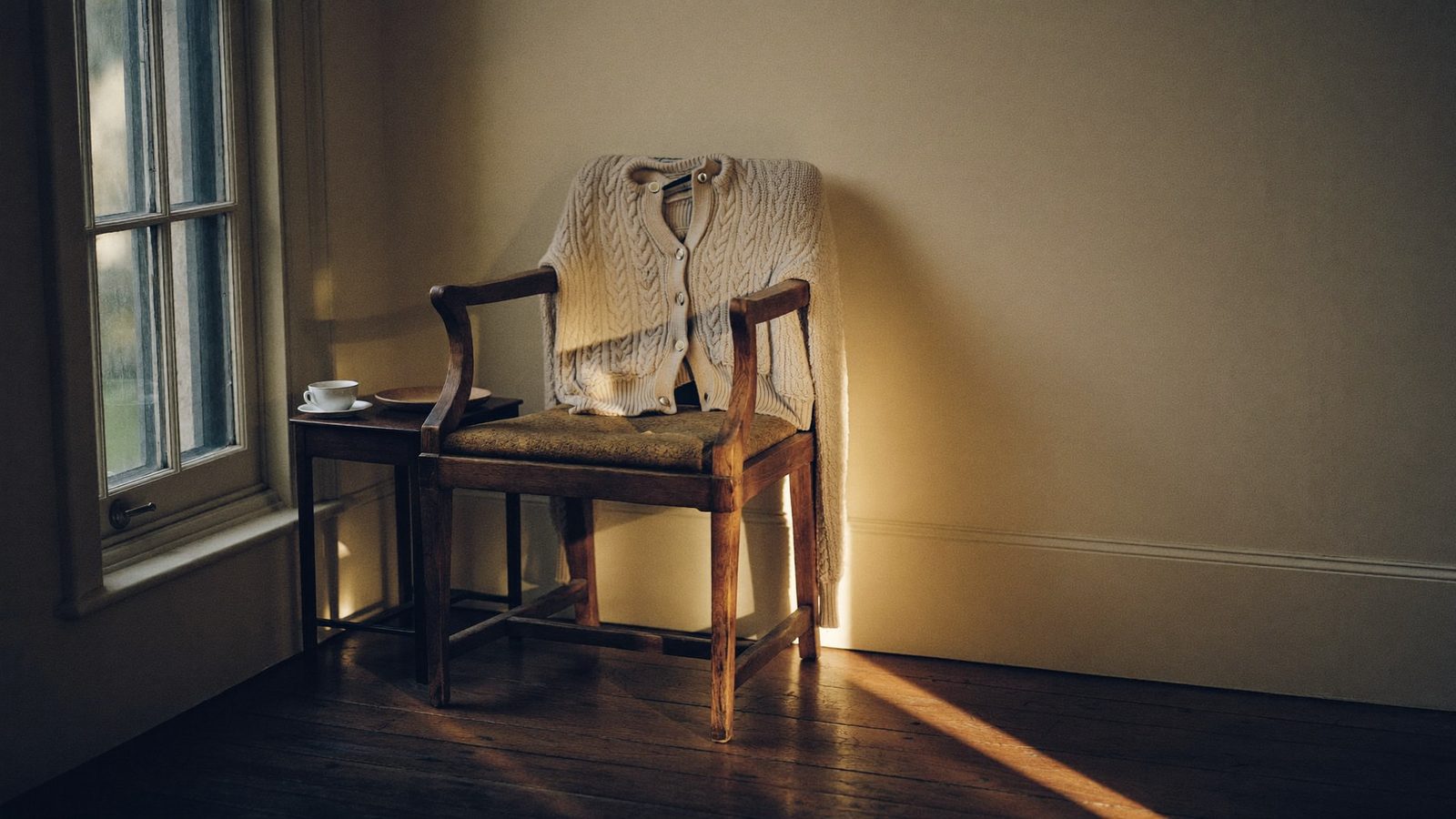 Ein leerer Stuhl an der Wand im späten Nachmittagslicht, eine Strickjacke über der Lehne, daneben ein Beistelltisch mit Tasse